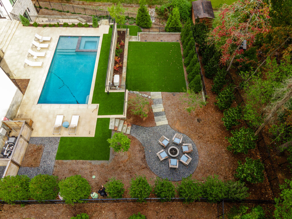 Aerial view of a modern backyard featuring a rectangular swimming pool with lounge chairs, artificial turf, a fire pit surrounded by seating, and neatly landscaped greenery offering privacy and structure.