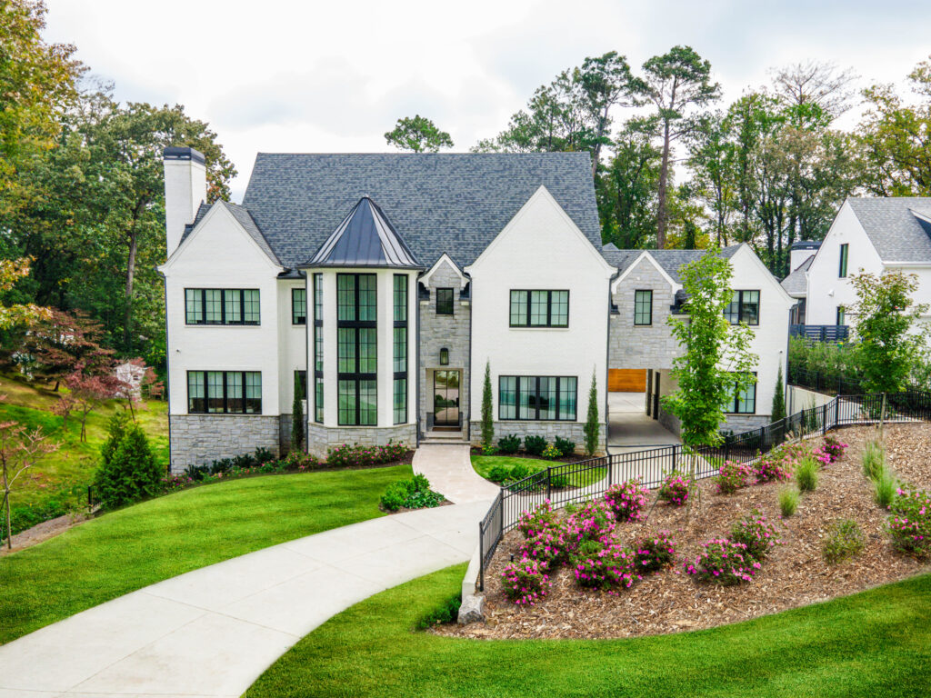 Modern white estate with black-framed windows and a turret-style entrance, set on a landscaped hill with a winding driveway, vibrant flowerbeds, and mature trees in the background.