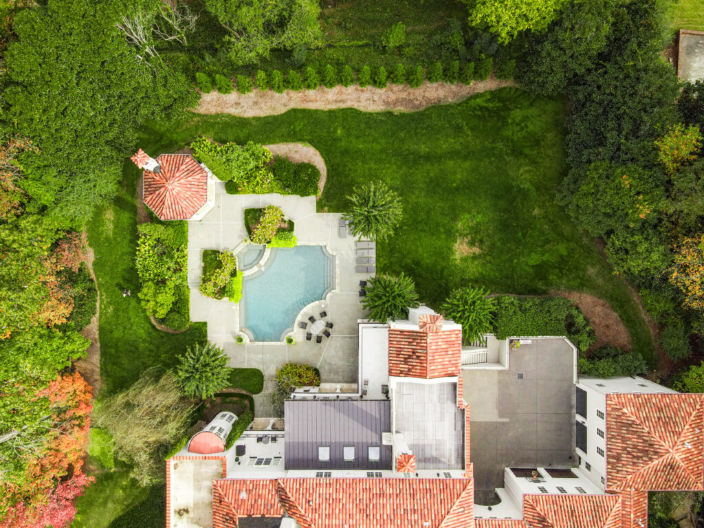 Aerial view of a luxury estate featuring a red-tile roof home, central swimming pool with surrounding patio, lush green lawn, and a charming gazebo nestled among mature trees and landscaped pathways.