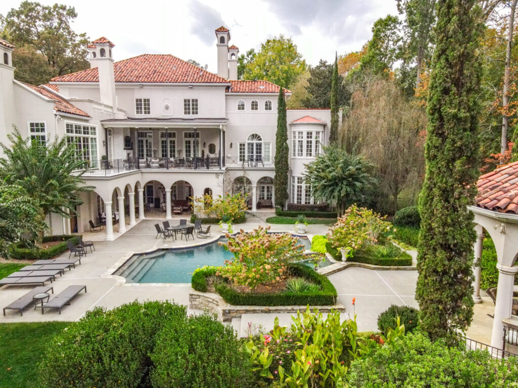 Luxurious estate with Mediterranean-style architecture, red-tiled roof, arched balconies, and a landscaped backyard featuring a swimming pool, lounge chairs, and manicured gardens surrounded by tall trees.