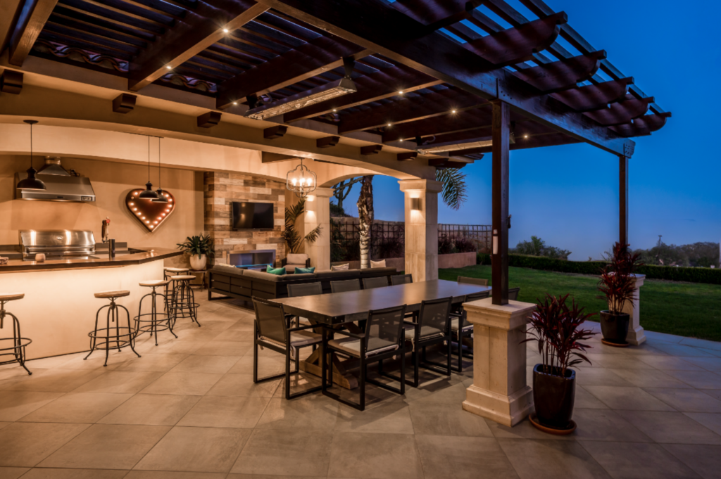 Elegant outdoor living space under a pergola at dusk, featuring a built-in kitchen with bar seating, a modern dining area, lounge furniture, a fireplace with a mounted TV, and ambient lighting.