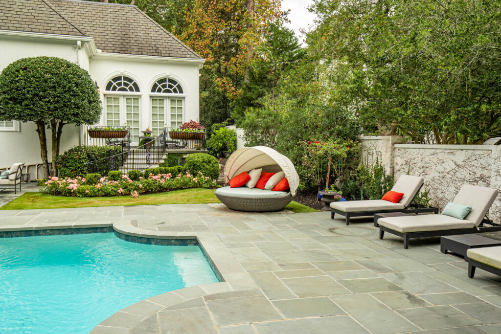 Elegant backyard with a stone patio, swimming pool, lounge chairs, and a round daybed with colorful pillows, surrounded by manicured hedges, flowering plants, and a classic white house with arched windows.