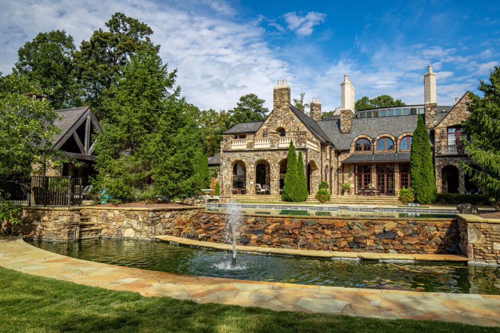 Luxurious estate with a grand stone mansion, infinity-edge pool, and tiered water feature, surrounded by lush greenery and manicured lawns, viewed from a golf putting green.