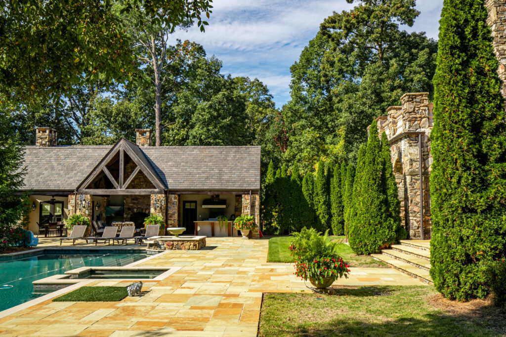 Luxury backyard with a stone guesthouse and covered outdoor lounge area, set beside a swimming pool and hot tub, surrounded by manicured hedges, potted plants, and tall evergreen trees.