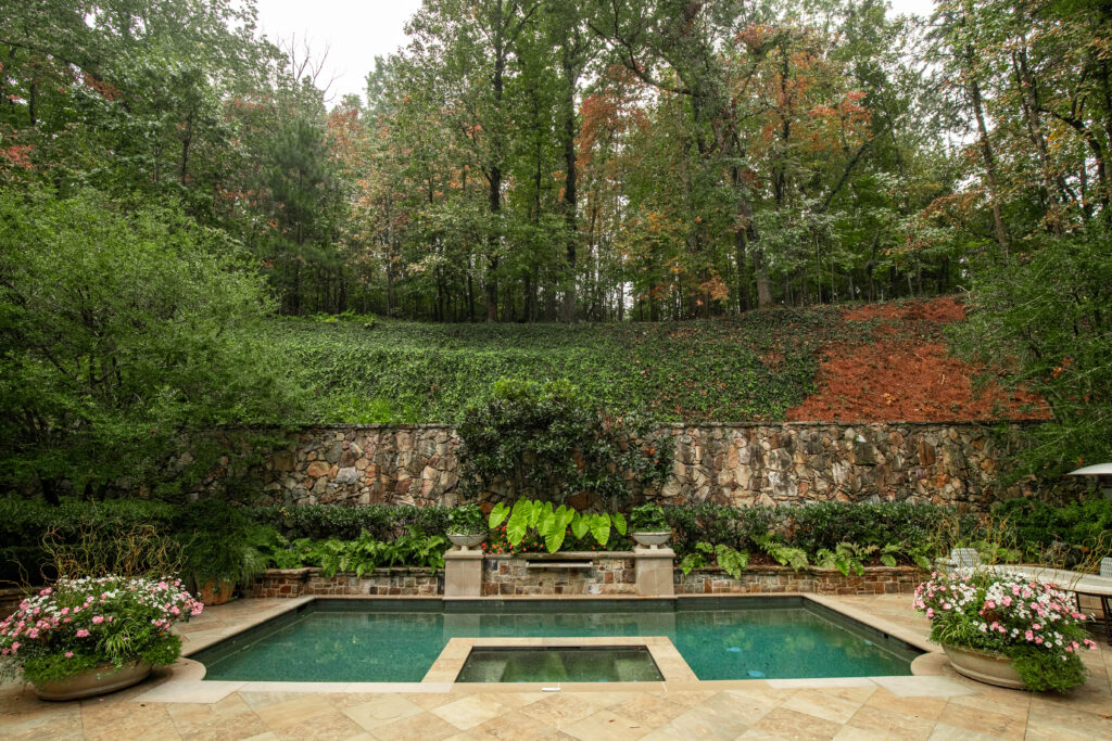 Secluded backyard pool with spa, surrounded by lush greenery, potted flowers, and a natural stone retaining wall set against a wooded hillside.
