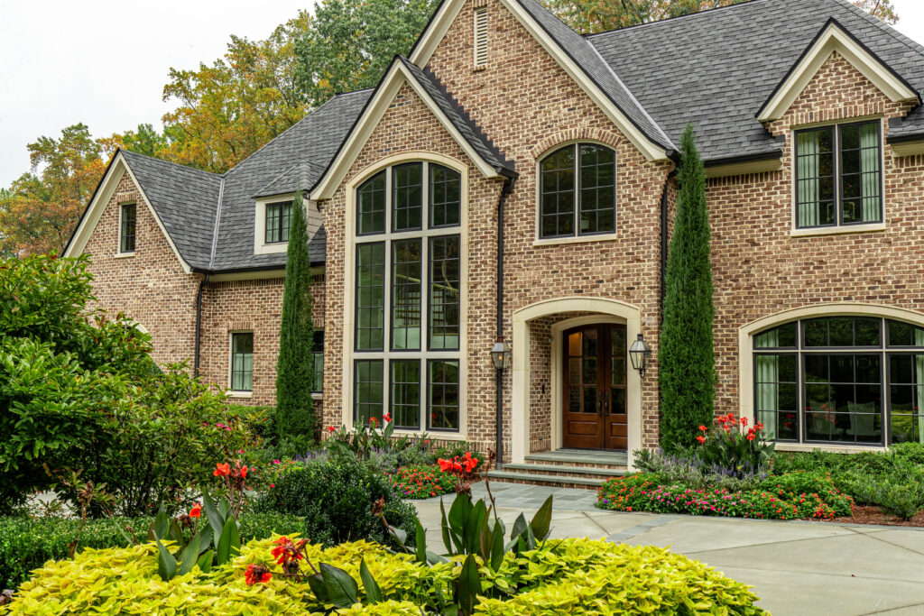 Beautiful brick home with tall arched windows, manicured garden beds, and a curved concrete walkway framed by colorful flowers, boxwoods, and ornamental trees.