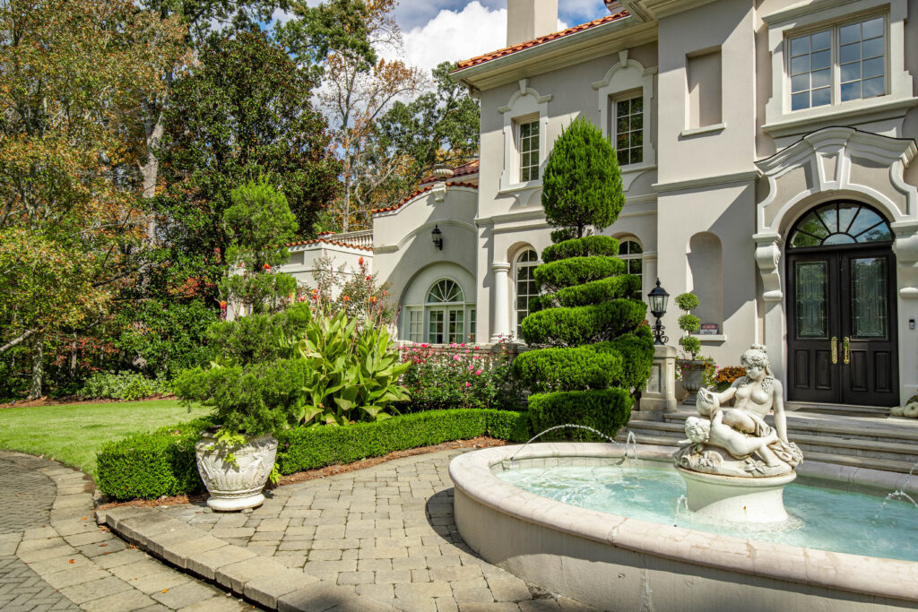 Elegant front yard of a classical estate featuring a circular fountain with a sculpted centerpiece, manicured hedges, topiary trees, and Mediterranean-inspired architecture.