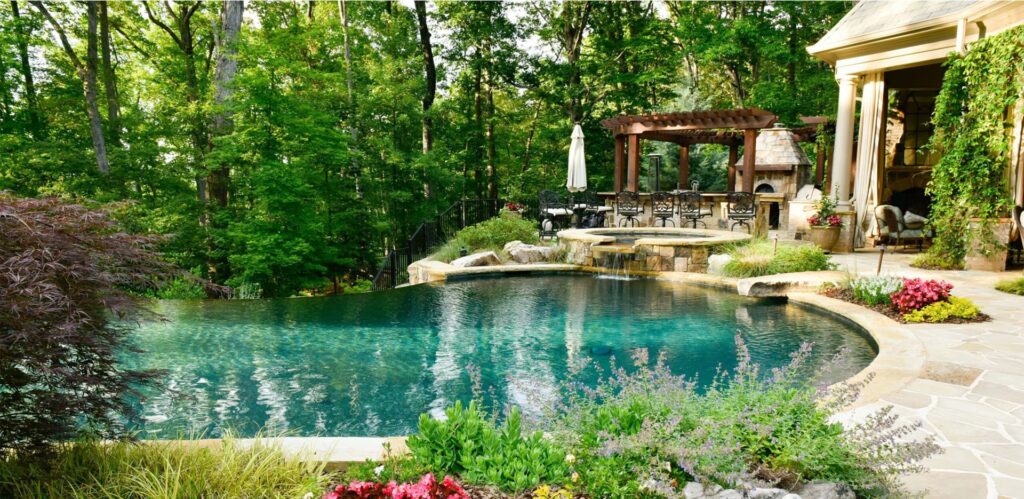 Luxurious backyard retreat with a lagoon-style infinity pool, elevated spa with a stone waterfall, outdoor bar under a pergola, and lush landscaping surrounded by tall forest trees.