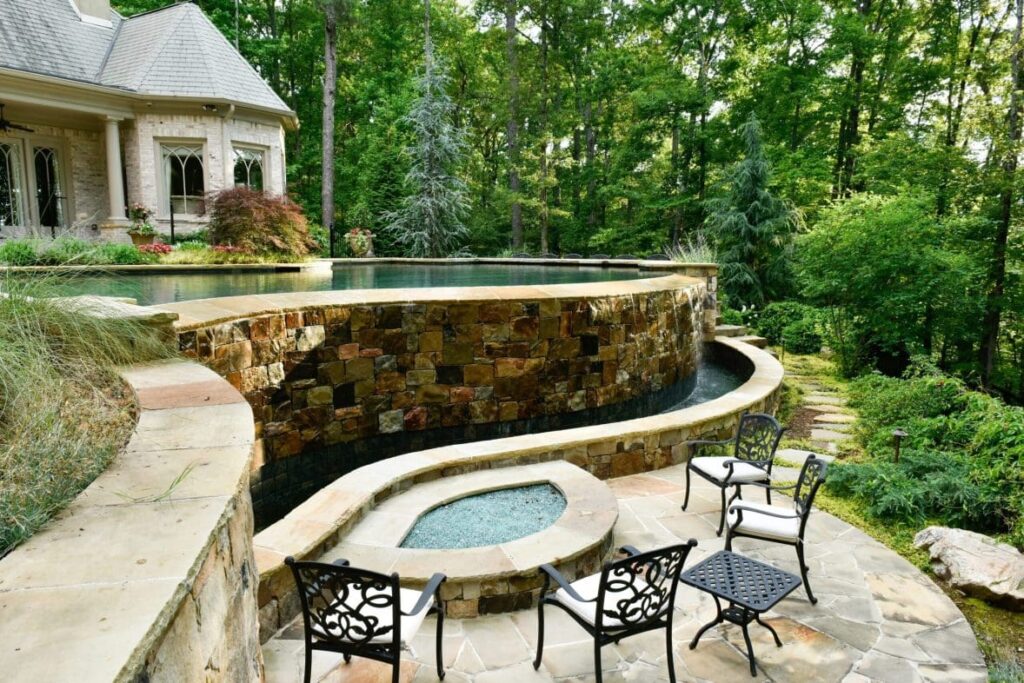 A stone infinity edge pool with a lower patio featuring iron seating and a small spa, surrounded by lush woodland landscaping.