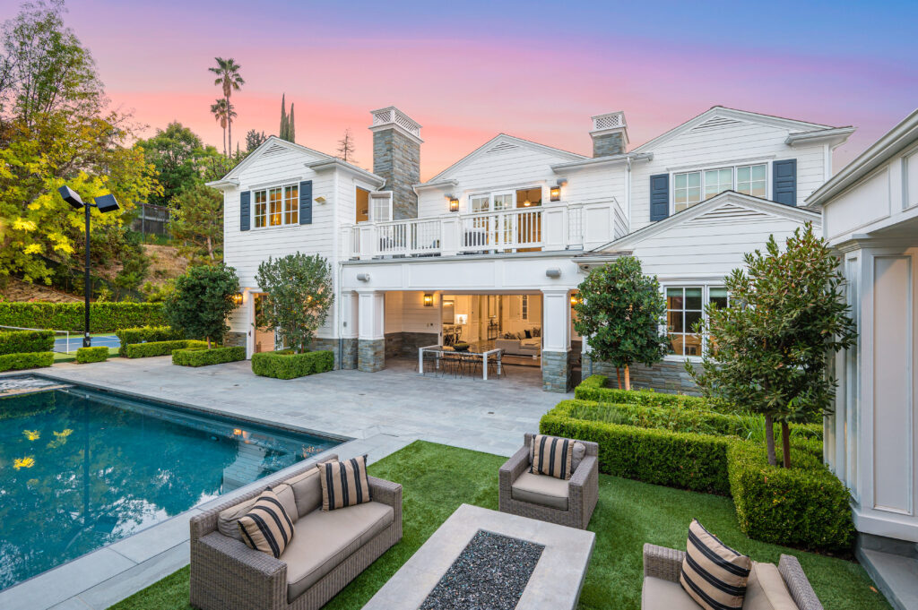 Elegant modern estate with white siding and dark shutters, featuring a large pool, fire pit seating area, and open-concept indoor-outdoor living space, set against a colorful sunset sky and lush landscaping.