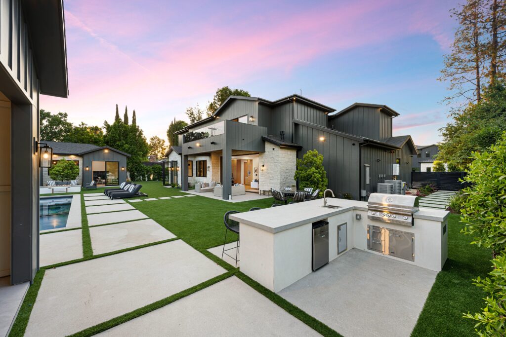 Modern backyard at sunset with a sleek outdoor kitchen and built-in grill, artificial turf, concrete stepping paths, lounge chairs by a pool, and a contemporary black and stone-clad home.