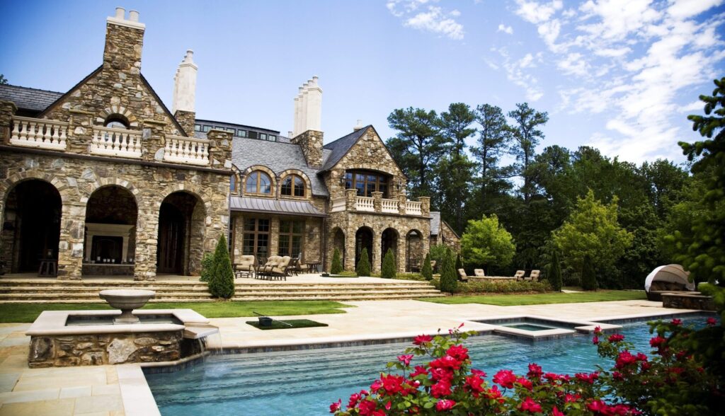 Luxury estate with rustic stone architecture, arched patio columns, and multiple chimneys overlooking a landscaped backyard with a pool, spa, and vibrant red flowers in bloom.
