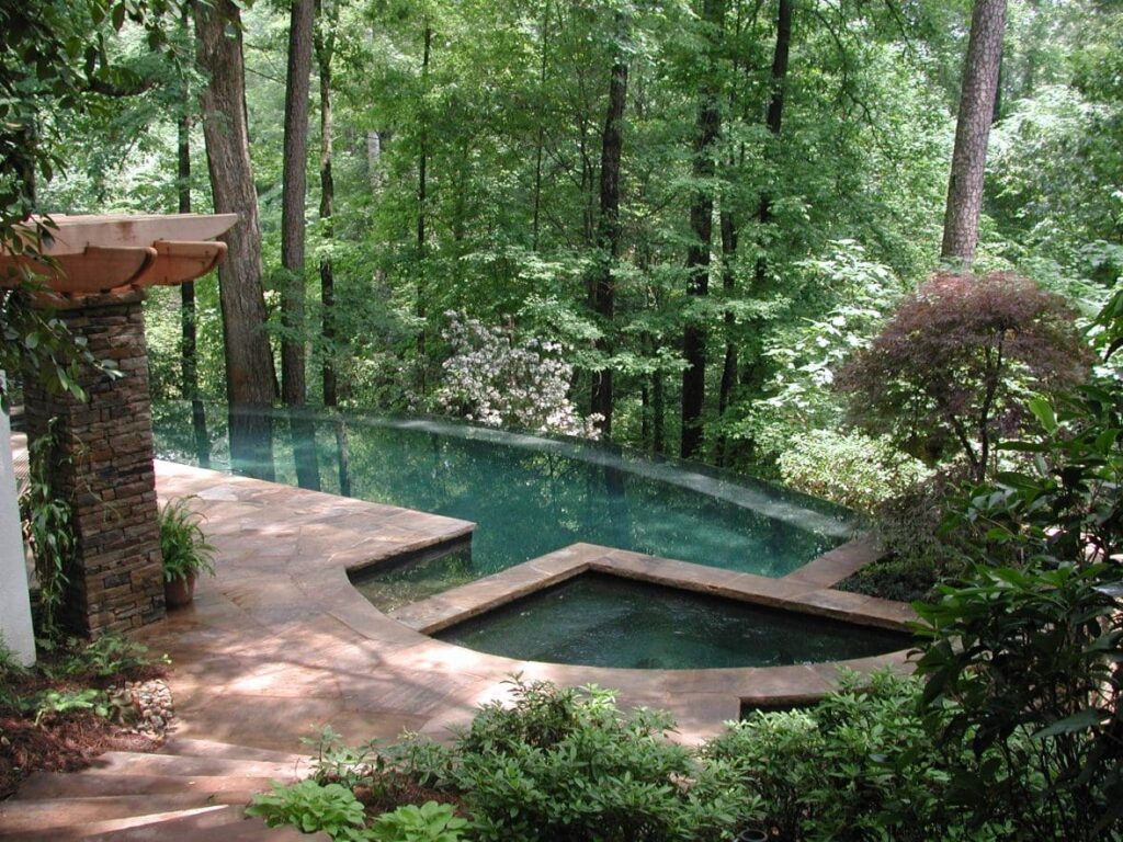 Infinity-edge pool surrounded by dense forest, featuring a geometric hot tub design and natural stone decking that blends seamlessly into the lush, wooded environment.