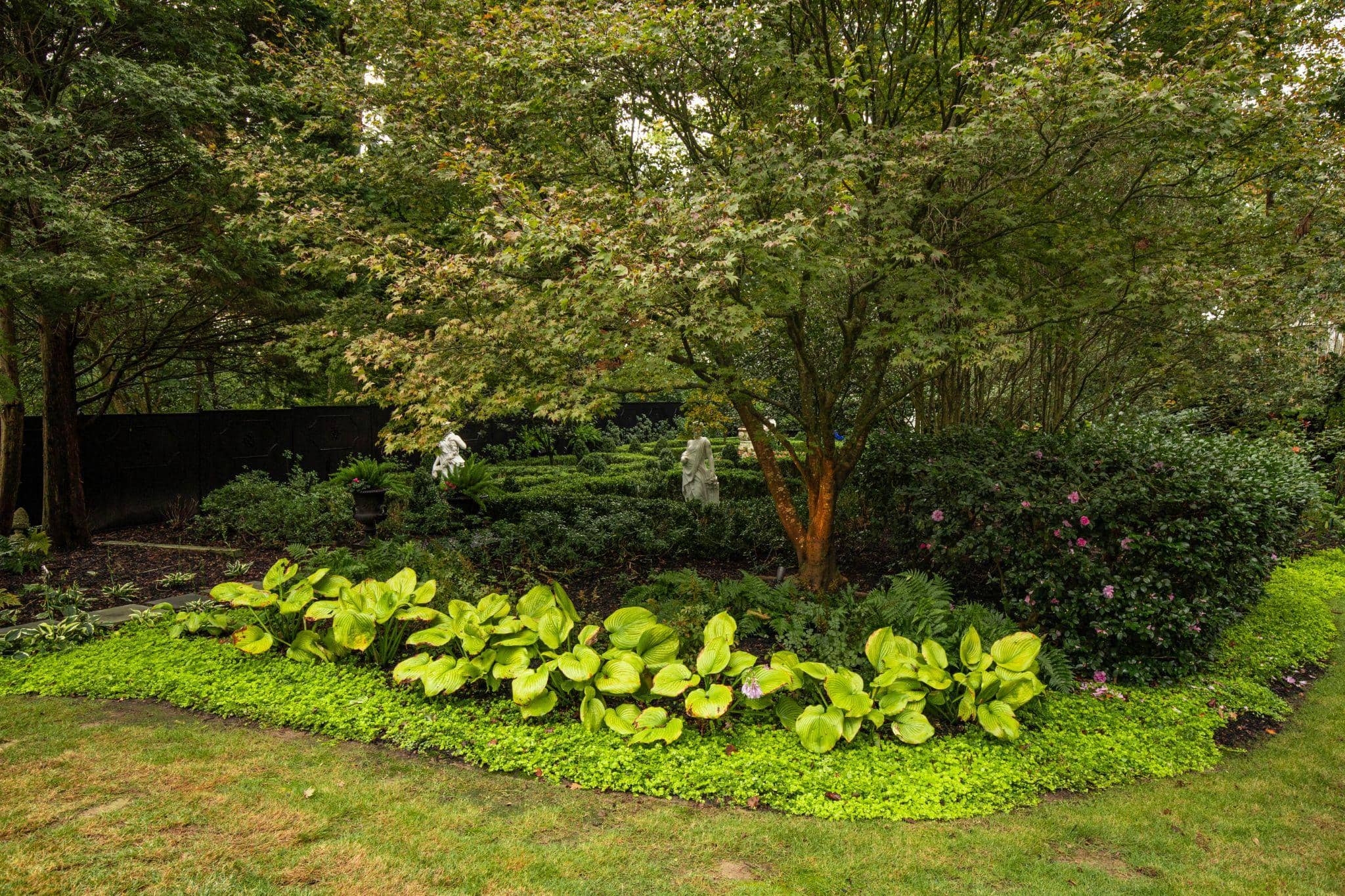 Lush shade garden with layered greenery, flowering shrubs, and classical statues